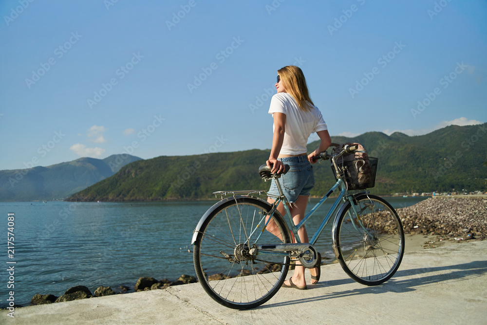 Young female wearing jeans shorts and white t-shirt standing with a bicycle by the sea and mountains 