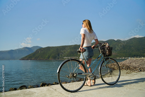 Young female wearing jeans shorts and white t-shirt standing with a bicycle by the sea and mountains 