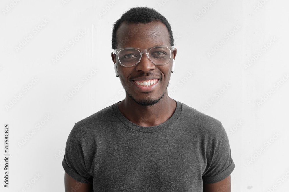 Horizontal shot of young dark-skinned man with positive smile on face, isolated on gray background, wearing plastic glasses, being in high spirit because of personal success, dressed in casual T-shirt Horizontal shot of young dark-skinned man with positive smile on face, isolated on gray background, wearing plastic glasses, being in high spirit because of personal success, dressed in casual T-shirt