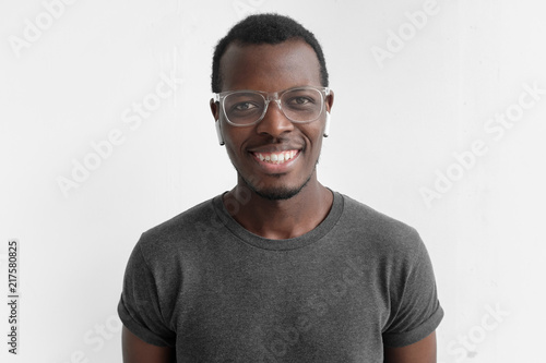 Horizontal shot of young dark-skinned man with positive smile on face, isolated on gray background, wearing plastic glasses, being in high spirit because of personal success, dressed in casual T-shirt