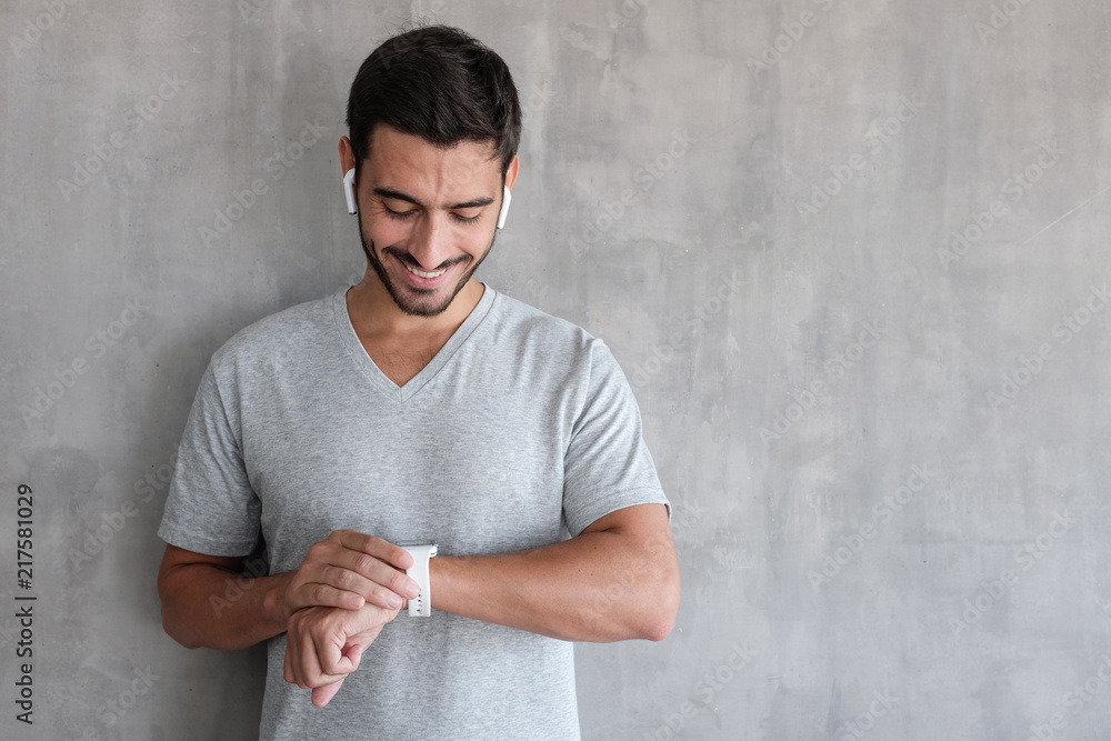 © Damir Khabirov - Young handsome man wearing wireless headphones and t shirt, checking smart watches with touch screen, standing against gray textured wall with copy space © Damir Khabirov - Young handsome man wearing wireless headphones and t shirt, checking smart watches with touch screen, standing against gray textured wall with copy space