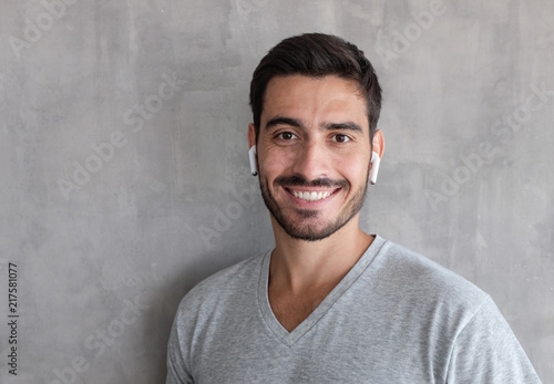 Close up headshot of young smiling handsome man wearing t shirt, listeting to his favorite music track, standing against gray textured wall