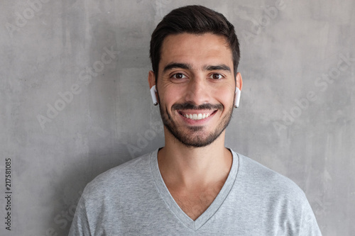 Young handsome man standing against gray textured wall, wearing t shirt and white wireless earphones, showing relaxed smile, looking confident and satisfied © Damir Khabirov