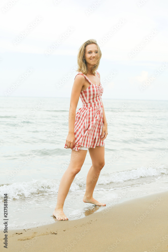 Young beautiful woman with blondie hair is laughing with toothy smile, wearing short red checked dress on seashore