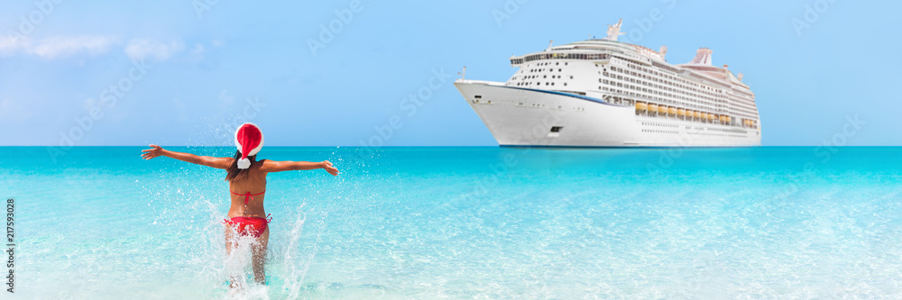 Christmas cruise travel vacation holidays in Caribbean beach. Happy bikini woman wearing santa claus hat on new year holiday running in happiness in blue turquoise water with ship in background.