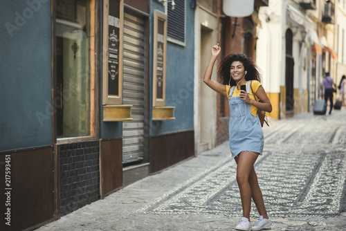 Young black woman with curly hair having fun in a urban surroundings. Mixed girl wearing cute casual clothes and earphones  smiling and walking happy while listening music in her smartphone.