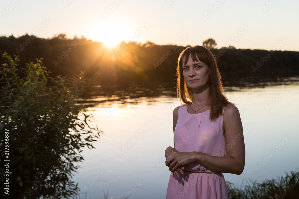 Girl in pink dress on the river beach during sunset. Outdoor portrait and sun background