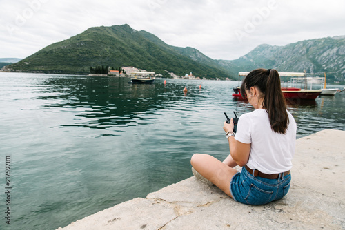 young adult woman sitting on pier with beautiful view of sea nd mountains. drone controller © phpetrunina14