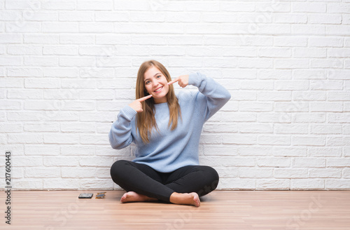 Young adult woman sitting on the floor in autumn over white brick wall smiling confident showing and pointing with fingers teeth and mouth. Health concept.