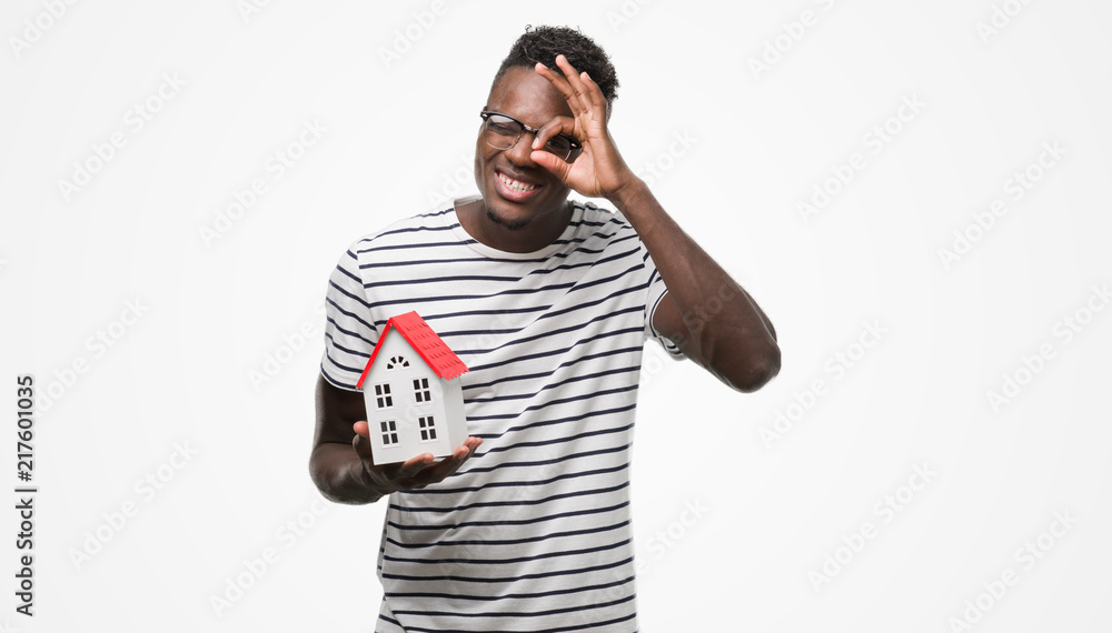 Young african american man holding house with happy face smiling doing ok sign with hand on eye looking through fingers Young african american man holding house with happy face smiling doing ok sign with hand on eye looking through fingers