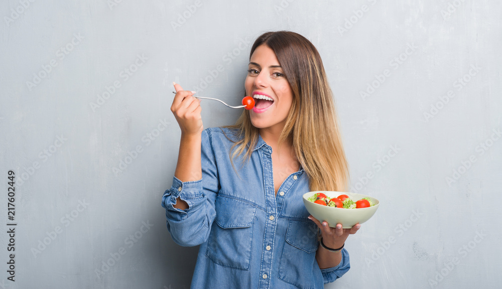 Young adult woman over grey grunge wall eating healthy tomato salad with a happy face standing and smiling with a confident smile showing teeth Young adult woman over grey grunge wall eating healthy tomato salad with a happy face standing and smiling with a confident smile showing teeth