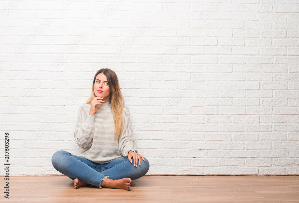 Young adult woman sitting on the floor over white brick wall at home with hand on chin thinking about question, pensive expression. Smiling with thoughtful face. Doubt concept. Young adult woman sitting on the floor over white brick wall at home with hand on chin thinking about question, pensive expression. Smiling with thoughtful face. Doubt concept.