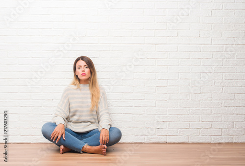Young adult woman sitting on the floor over white brick wall at home with serious expression on face. Simple and natural looking at the camera.