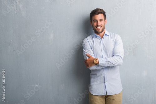 Handsome young business man over grey grunge wall wearing elegant shirt happy face smiling with crossed arms looking at the camera. Positive person.