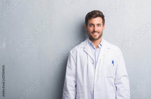 Foto Handsome young professional man over grey grunge wall wearing white coat with a happy and cool smile on face