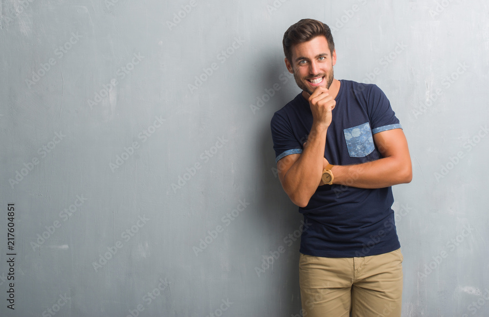 Handsome young man over grey grunge wall looking confident at the camera with smile with crossed arms and hand raised on chin. Thinking positive. Handsome young man over grey grunge wall looking confident at the camera with smile with crossed arms and hand raised on chin. Thinking positive.