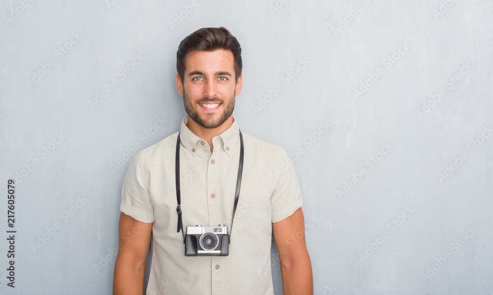 Handsome young man over grey grunge wall holding vintage photo camera with a happy face standing and smiling with a confident smile showing teeth Handsome young man over grey grunge wall holding vintage photo camera with a happy face standing and smiling with a confident smile showing teeth