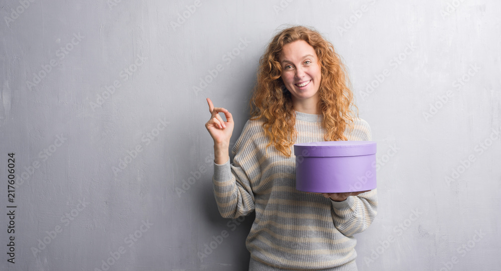 Young redhead woman over grey grunge wall holding box very happy pointing with hand and finger to the side Young redhead woman over grey grunge wall holding box very happy pointing with hand and finger to the side