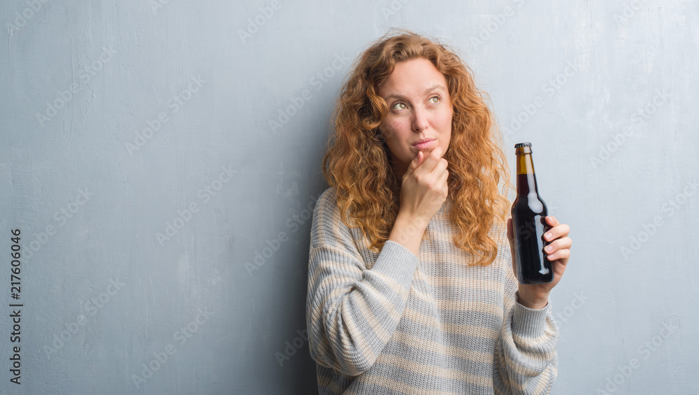 Young redhead woman over grey grunge wall holding beer bottle serious face thinking about question, very confused idea Young redhead woman over grey grunge wall holding beer bottle serious face thinking about question, very confused idea