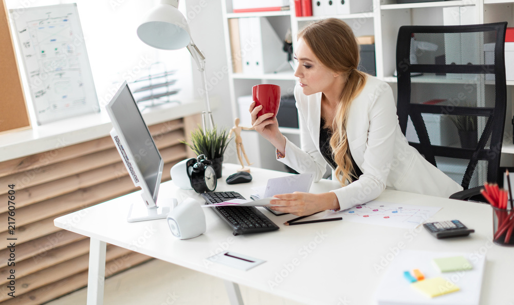 A young girl is sitting at the computer desk in the office and is holding a cup and documents. Before the girl on the table are diagrams. A young girl is sitting at the computer desk in the office and is holding a cup and documents. Before the girl on the table are diagrams.