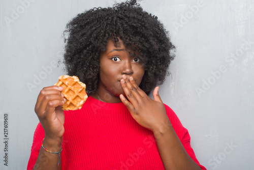 Young african american woman over grey grunge wall eating belgium waffle cover mouth with hand shocked with shame for mistake, expression of fear, scared in silence, secret concept