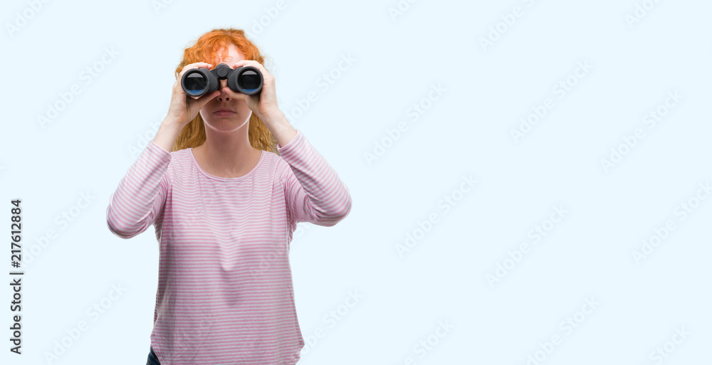 Young redhead woman looking through binoculars with a confident expression on smart face thinking serious