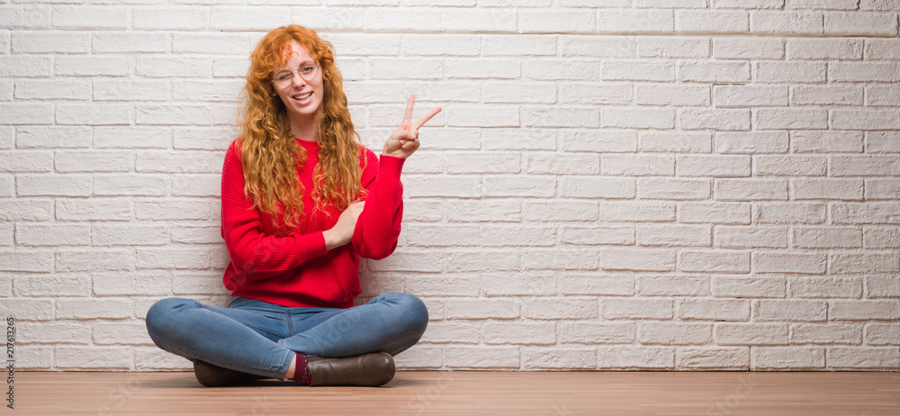 Young redhead woman sitting over brick wall smiling with happy face winking at the camera doing victory sign. Number two. Young redhead woman sitting over brick wall smiling with happy face winking at the camera doing victory sign. Number two.