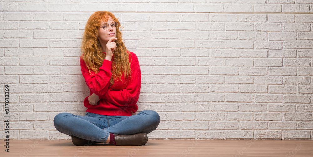 Young redhead woman sitting over brick wall looking confident at the camera with smile with crossed arms and hand raised on chin. Thinking positive. Young redhead woman sitting over brick wall looking confident at the camera with smile with crossed arms and hand raised on chin. Thinking positive.