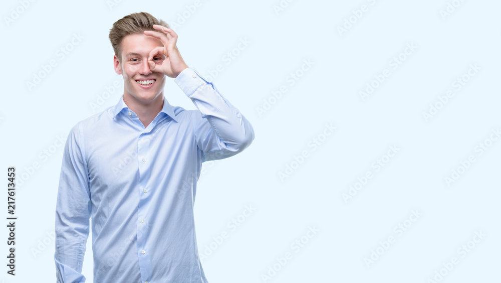 Young handsome blond man with happy face smiling doing ok sign with hand on eye looking through fingers Young handsome blond man with happy face smiling doing ok sign with hand on eye looking through fingers
