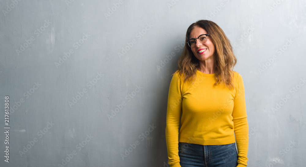 Fototapeta premium Middle age hispanic woman over grey wall wearing glasses looking away to side with smile on face, natural expression. Laughing confident.