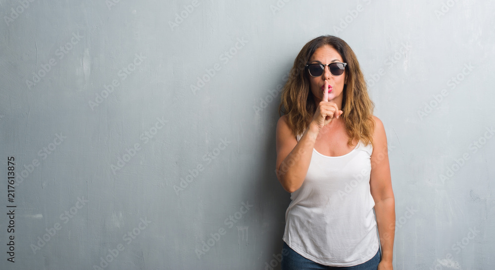 Middle age hispanic woman over grey wall wearing sunglasses asking to be quiet with finger on lips. Silence and secret concept. Middle age hispanic woman over grey wall wearing sunglasses asking to be quiet with finger on lips. Silence and secret concept.