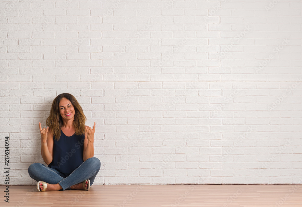 Middle age hispanic woman sitting on the floor over white brick wall shouting with crazy expression doing rock symbol with hands up. Music star. Heavy concept. Middle age hispanic woman sitting on the floor over white brick wall shouting with crazy expression doing rock symbol with hands up. Music star. Heavy concept.