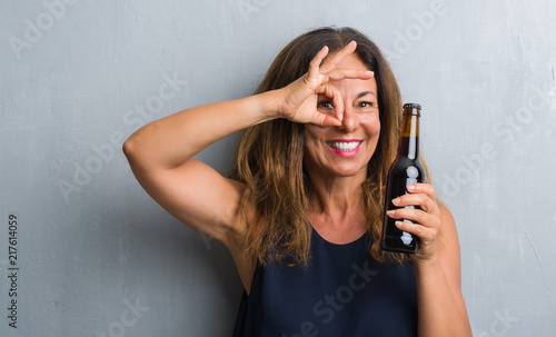 Middle age hispanic woman standing over grey grunge wall holding beer bottle with happy face smiling doing ok sign with hand on eye looking through fingers