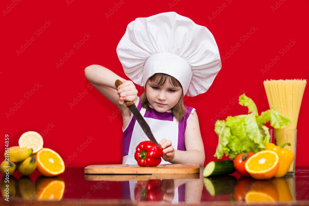 Happy little girl in chef uniform cuts vegetables in kitchen. Kid chef. Vegetarian. Cooking Process Concept Happy little girl in chef uniform cuts vegetables in kitchen. Kid chef. Vegetarian. Cooking Process Concept