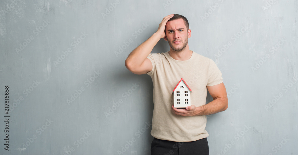 Young caucasian real state agent man over grey grunge wall holding house stressed with hand on head, shocked with shame and surprise face, angry and frustrated. Fear and upset for mistake. Young caucasian real state agent man over grey grunge wall holding house stressed with hand on head, shocked with shame and surprise face, angry and frustrated. Fear and upset for mistake.