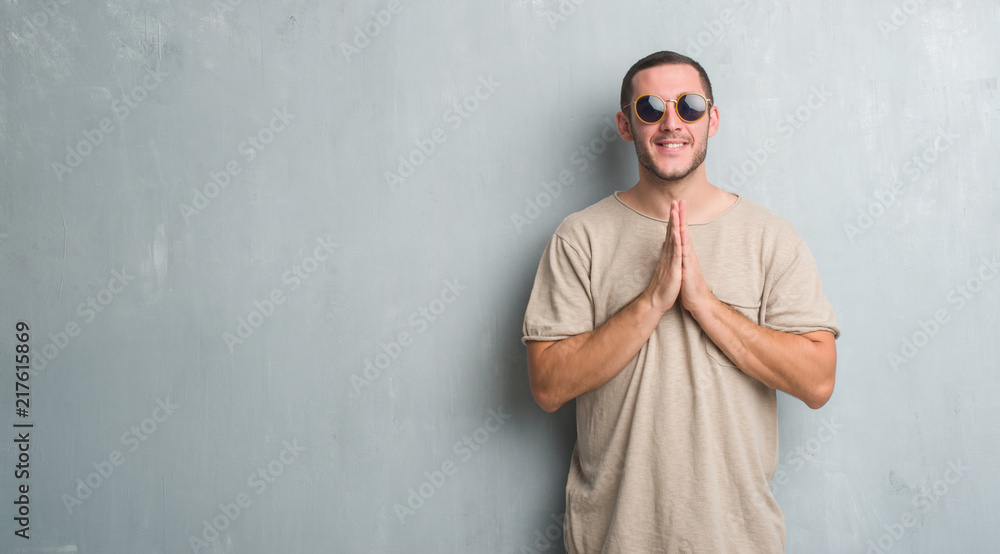 Young caucasian man over grey grunge wall wearing sunglasses praying with hands together asking for forgiveness smiling confident. Young caucasian man over grey grunge wall wearing sunglasses praying with hands together asking for forgiveness smiling confident.