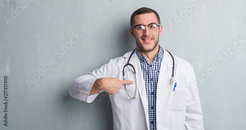 Young caucasian man over grey grunge wall wearing doctor uniform with surprise face pointing finger to himself