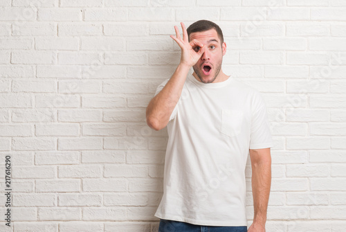 Young caucasian man standing over white brick wall doing ok gesture like binoculars sticking tongue out, eyes looking through fingers. Crazy expression.