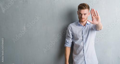 Young redhead business man over grey grunge wall doing stop sing with palm of the hand. Warning expression with negative and serious gesture on the face.