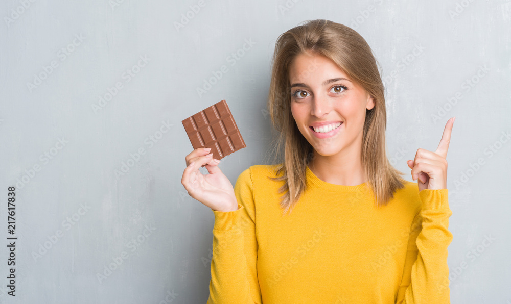 Beautiful young woman over grunge grey wall eating chocolate bar surprised with an idea or question pointing finger with happy face, number one Beautiful young woman over grunge grey wall eating chocolate bar surprised with an idea or question pointing finger with happy face, number one