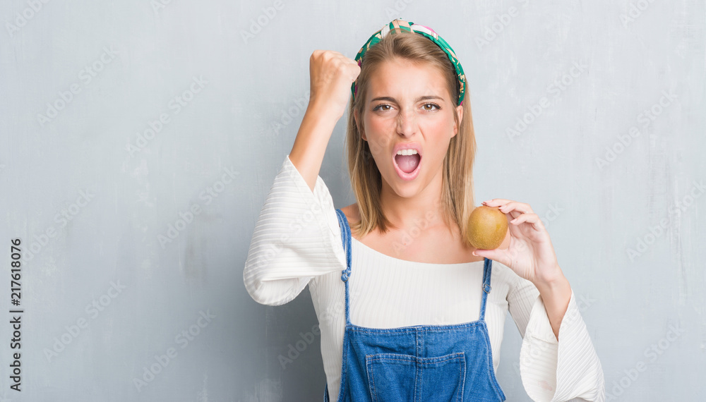 Beautiful young woman over grunge grey wall holding fresh kiwi annoyed and frustrated shouting with anger, crazy and yelling with raised hand, anger concept Beautiful young woman over grunge grey wall holding fresh kiwi annoyed and frustrated shouting with anger, crazy and yelling with raised hand, anger concept