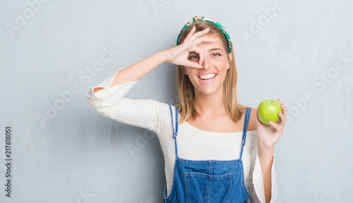 Beautiful young woman over grunge grey wall eating green apple with happy face smiling doing ok sign with hand on eye looking through fingers