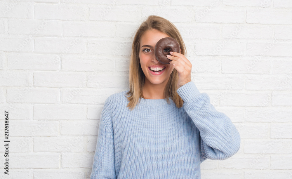 Beautiful young woman over white brick wall eating chocolate donut with a happy face standing and smiling with a confident smile showing teeth Beautiful young woman over white brick wall eating chocolate donut with a happy face standing and smiling with a confident smile showing teeth