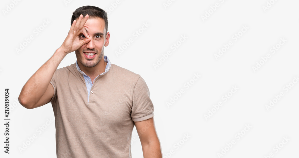 Handsome young man with happy face smiling doing ok sign with hand on eye looking through fingers Handsome young man with happy face smiling doing ok sign with hand on eye looking through fingers