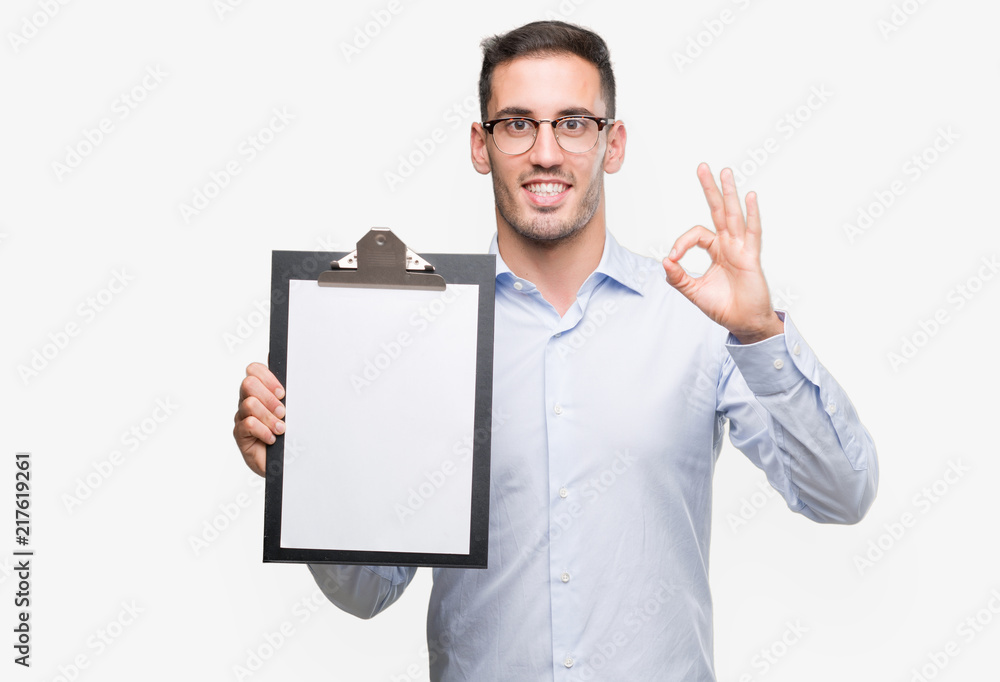 Handsome young business man showing a clipboard doing ok sign with fingers, excellent symbol Handsome young business man showing a clipboard doing ok sign with fingers, excellent symbol