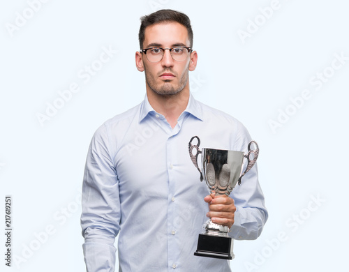 Handsome young man holding trophy with a confident expression on smart face thinking serious