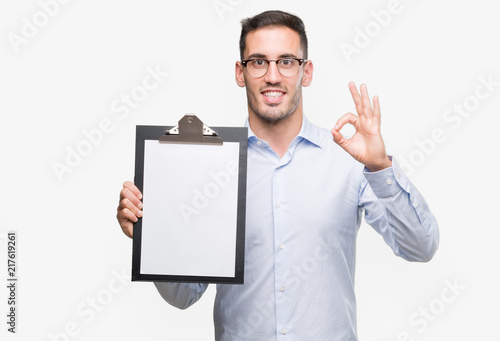 Handsome young business man showing a clipboard doing ok sign with fingers, excellent symbol