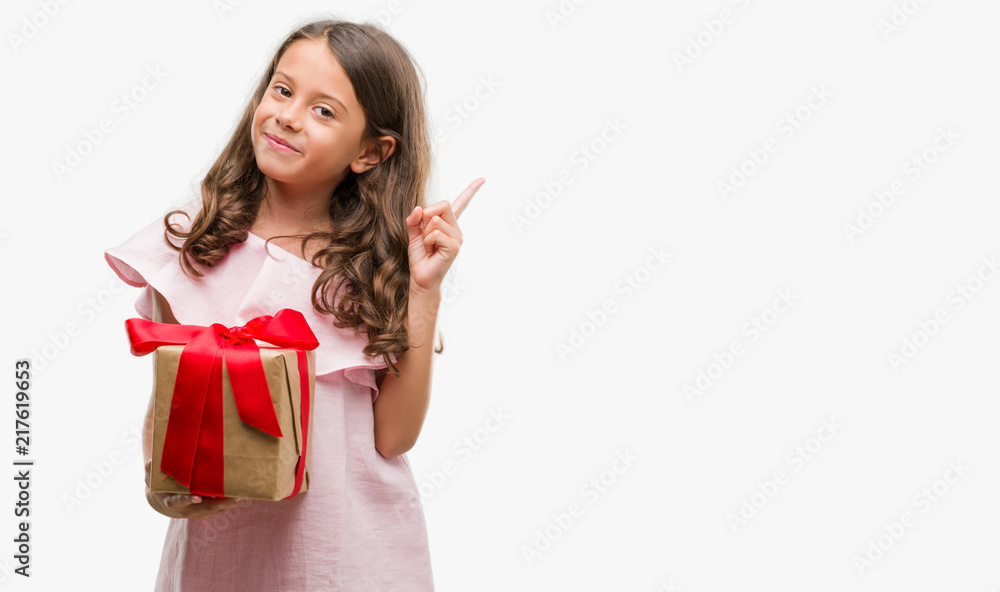 Brunette hispanic girl holding a gift very happy pointing with hand and finger to the side Brunette hispanic girl holding a gift very happy pointing with hand and finger to the side