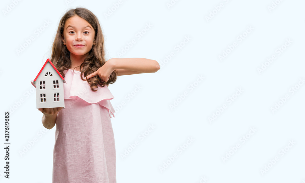 Brunette hispanic girl holding little house with surprise face pointing finger to himself Brunette hispanic girl holding little house with surprise face pointing finger to himself