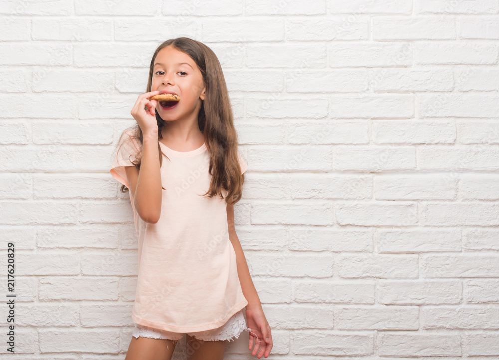 Young hispanic kid over white brick wall eating chocolate chips cooky with a confident expression on smart face thinking serious Young hispanic kid over white brick wall eating chocolate chips cooky with a confident expression on smart face thinking serious